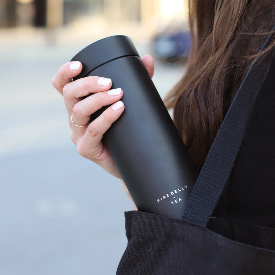 Person holding a black travel tea tumbler labeled 'Firebelly Tea.'